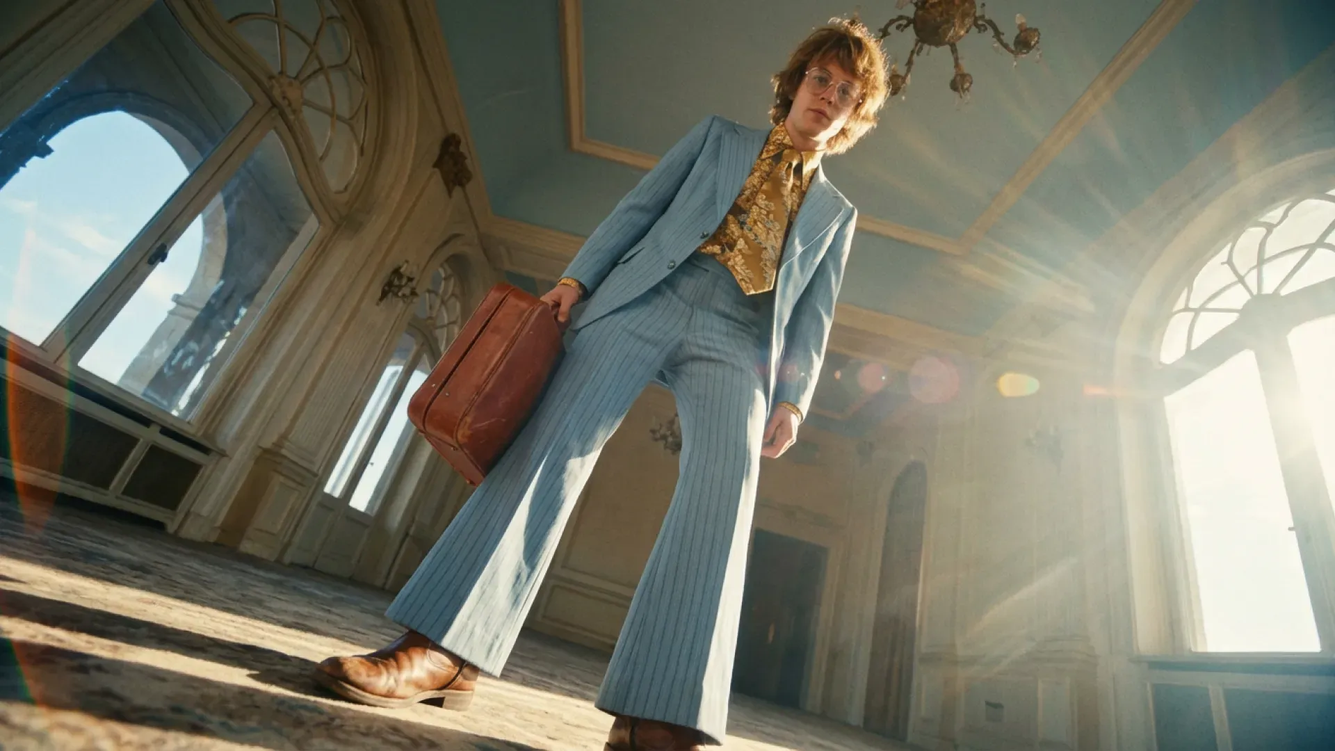 Low-angle fashion portrait of a person in a pale blue suit carrying a brown bag in a sunlit hall